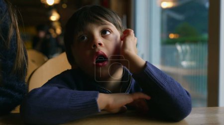 Photo for Bored small boy yawning while waiting at restaurant table by window. Child feeling boredom in authentic lifestyle moment awaiting for food to arrive - Royalty Free Image