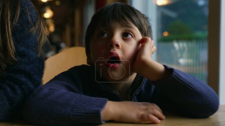 Photo for Bored small boy yawning while waiting at restaurant table by window. Child feeling boredom in authentic lifestyle moment awaiting for food to arrive - Royalty Free Image