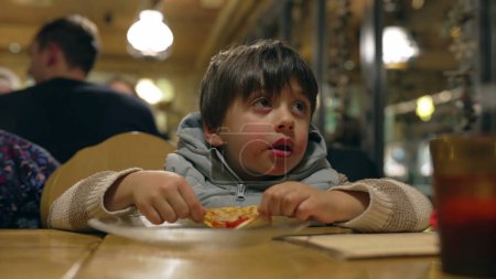 Foto de Niño pequeño comiendo rebanada de pizza en el restaurante - Niño disfrutando de la comida rica en carbohidratos en el comedor por la noche - Imagen libre de derechos