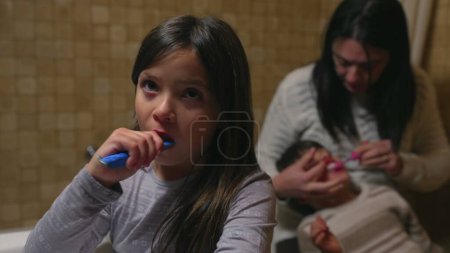 Foto de Momento de cuidado dental familiar - Madre asistiendo al hijo mientras su hija cepilla los dientes por el fregadero del baño - Imagen libre de derechos