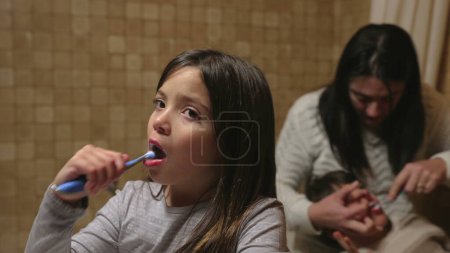 Téléchargez les photos : Scène d'hygiène dentaire familiale - Mère aide le jeune enfant à se brosser les dents pendant que sa sœur aînée se brosse les dents dans la salle de bain au premier plan - en image libre de droit