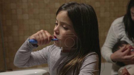 Foto de El niño cepilla los dientes frente al espejo del baño, mientras que la madre ayuda a los hermanos más pequeños con la higiene dental en el fondo - Imagen libre de derechos