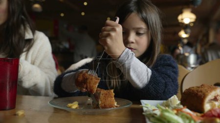 Téléchargez les photos : Petite fille mangeant de la nourriture au restaurant, enfant dégustant du poulet cordon bleu pour les repas à l'intérieur en bois confortable. Famille au dîner - en image libre de droit