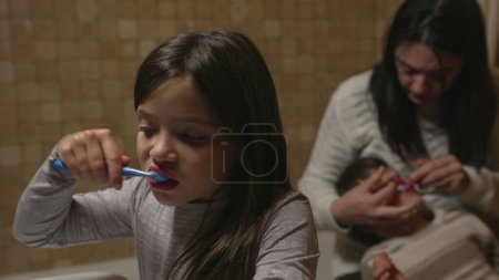 Foto de El niño cepilla los dientes frente al espejo del baño, mientras que la madre ayuda a los hermanos más pequeños con la higiene dental en el fondo - Imagen libre de derechos