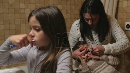 Foto de Escena de higiene dental familiar: la madre ayuda al niño más pequeño mientras la hermana mayor cepilla los dientes en el baño en primer plano - Imagen libre de derechos
