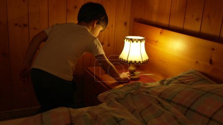 Foto de Un niño pequeño apaga la lámpara de noche en el interior acogedor interior de madera. Decoración tradicional en un ambiente hogareño - Imagen libre de derechos