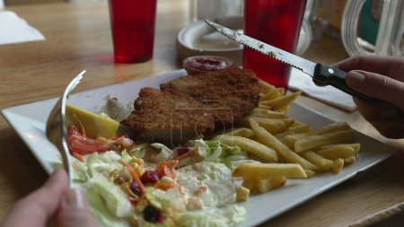 Téléchargez les photos : Gros plan de assiette de viande milanaise avec salade et frites. Couper les aliments à la main avec un couteau - déjeuner - en image libre de droit