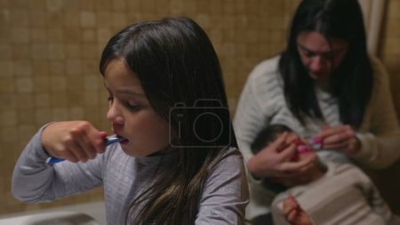 Foto de Momento de cuidado dental familiar - Madre asistiendo al hijo mientras su hija cepilla los dientes por el fregadero del baño - Imagen libre de derechos