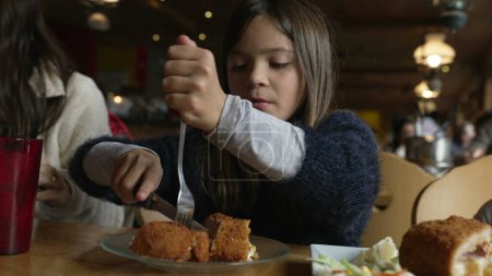Téléchargez les photos : Petite fille mangeant de la nourriture au restaurant, enfant dégustant du poulet cordon bleu pour les repas à l'intérieur en bois confortable. Famille au dîner - en image libre de droit