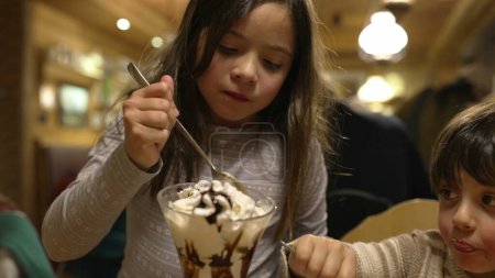 Foto de Hermanos compartiendo helado con crema batida en restaurante Diner - Niños disfrutando del postre de azúcar Tratar Indulgencia - Imagen libre de derechos