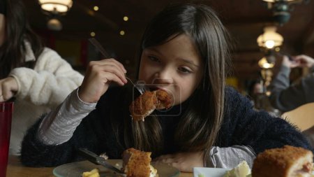 Foto de Niña comiendo comida en el restaurante, niño disfrutando de cordon bleu pollo para la hora de comer en el acogedor interior de madera. Familia en el restaurante - Imagen libre de derechos