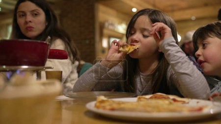 Photo for Small girl eating slice of pizza at restaurant, family enjoying time together during weekend activity at evening diner - Royalty Free Image