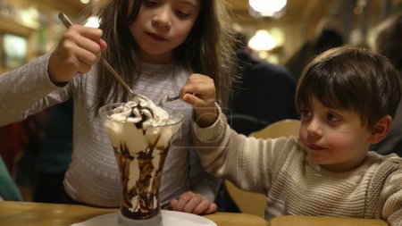 Foto de Hermanos compartiendo helado con crema batida en restaurante Diner - Niños disfrutando del postre de azúcar Tratar Indulgencia - Imagen libre de derechos