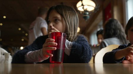Téléchargez les photos : Petite fille buvant de l'eau de la tasse rouge en plastique au restaurant en attendant que la nourriture arrive, dîner intérieur en bois à côté de frère frère frère - en image libre de droit