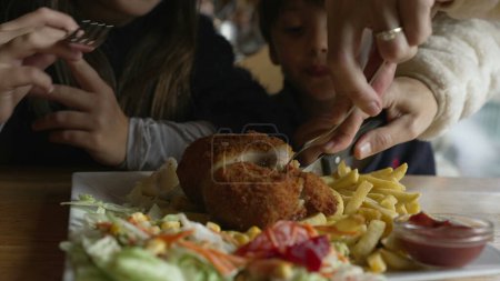 Photo for Children at restaurant about to eat cordon bleu food mealtime, closeup parent hand cutting meat, preparing for son and daughter. Little boy grabbing fry with ketchup - Royalty Free Image