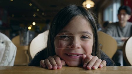 Photo for Closeup face of cute little girl leaning on table at restaurant, looking at camera smiling. Adorable child feeling happy and laughing - Royalty Free Image