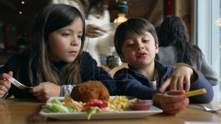 Foto de Niños comiendo comida en el restaurante, hermanos: hermano pequeño y hermana disfrutando de papas fritas y comida de pollo en el comedor dentro del interior de madera - Imagen libre de derechos