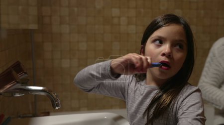 Téléchargez les photos : Fille brossant les dents devant le miroir de salle de bain par évier tandis que la mère aide le petit frère à brosser les dents, hygiène dentaire familiale authentique - en image libre de droit