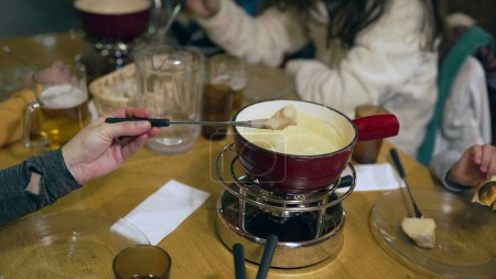 Foto de Primer plano de fondue tradicional suiza en la mesa del restaurante, olla roja con calentador - sumergiendo pedazo de pan dentro de queso derretido. La gente disfruta de la comida en Suiza - Imagen libre de derechos