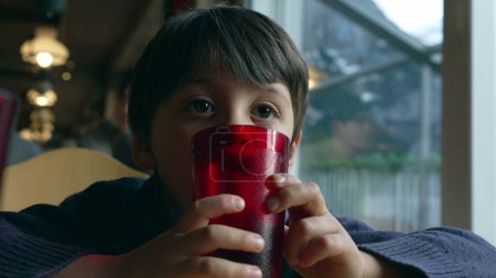 Téléchargez les photos : Pensif petit garçon rêvant au restaurant appuyé sur une tasse en plastique rouge tout en regardant à blanc, enfant réfléchi attendant la nourriture au dîner par la fenêtre - en image libre de droit