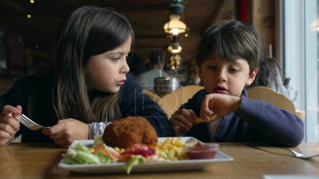 Foto de Niños comiendo comida en el restaurante, hermanos: hermano pequeño y hermana disfrutando de papas fritas y comida de pollo en el comedor dentro del interior de madera - Imagen libre de derechos