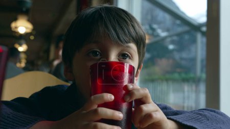 Téléchargez les photos : Pensif petit garçon rêvant au restaurant appuyé sur une tasse en plastique rouge tout en regardant à blanc, enfant réfléchi attendant la nourriture au dîner par la fenêtre - en image libre de droit
