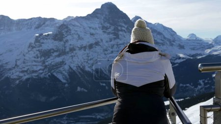 Téléchargez les photos : Dos de la personne contemplative debout dehors pendant la journée d'hiver surplombant de belles montagnes pittoresques dans les Alpes, femme coûteuse portant bonnet et veste - en image libre de droit