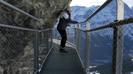 Foto de Mujer turista cautelosa caminando por el acantilado sosteniendo la sensación de barandilla de metal prudente sobre la altura elevada con paisaje montañoso escénico en los Alpes suizos - Imagen libre de derechos