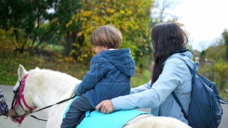 Téléchargez les photos : Enfant monte à cheval poney, mère souriante et petit garçon fils pendant la saison d'automne - en image libre de droit