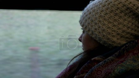 Téléchargez les photos : Contemplation du voyage en train - Femme réfléchie en bonnet et écharpe, regardant passer le paysage par la fenêtre du train pendant l'hiver - en image libre de droit