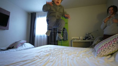 Téléchargez les photos : Heureux enfant sautant de haut en bas dans le lit de l'hôtel. Petit garçon insouciant rebondissant - en image libre de droit