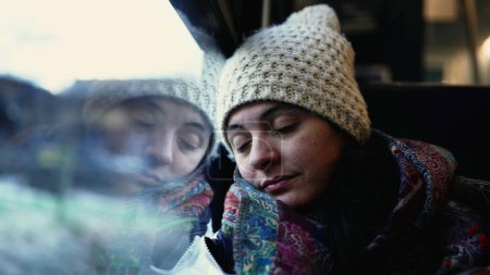 Téléchargez les photos : Passager endormi dans le train avec vue sur le paysage. Femme penche la tête sur la fenêtre voyageant pendant la saison d'hiver, portant l'écharpe et le bonnet - en image libre de droit
