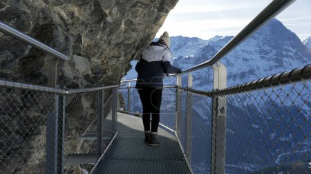 Foto de Mujer turista cautelosa caminando por el acantilado sosteniendo la sensación de barandilla de metal prudente sobre la altura elevada con paisaje montañoso escénico en los Alpes suizos - Imagen libre de derechos