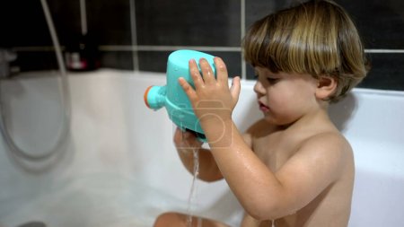 Téléchargez les photos : Petit garçon à l'intérieur de la baignoire jouant avec un objet jouet en plastique, un garçon caucasien vu à travers le verre pendant la routine de bain nocturne - en image libre de droit