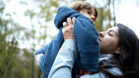 Photo for Mother lifting on in arms and kissing in cheek standing outside during autumn fall season, authentic lifestyle family scene of mom and child loving relationship - Royalty Free Image