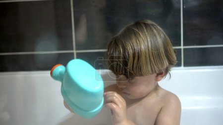 Téléchargez les photos : Petit garçon à l'intérieur de la baignoire jouant avec un objet jouet en plastique, un garçon caucasien vu à travers le verre pendant la routine de bain nocturne - en image libre de droit