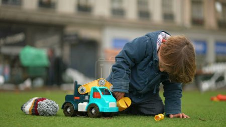 Téléchargez les photos : Petit garçon jouant avec camion à l'extérieur au parc pendant la journée d'automne. Bonnet et veste enfant immergés dans le jeu de l'objet - en image libre de droit