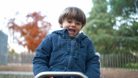 Photo for Joyful child plays at park on top of seesaw, Active 3 year old boy kid wearing blue jacket enjoys autumn day outdoors - Royalty Free Image
