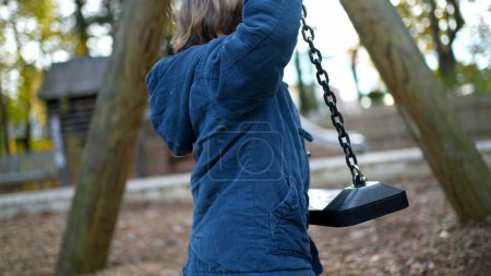 Foto de Niño tratando de escalar el columpio del parque durante el día de otoño usando chaqueta azul. Un pequeño niño de 3 años intenta sentarse en la cima del columpio - prueba y error concepto de la infancia - Imagen libre de derechos