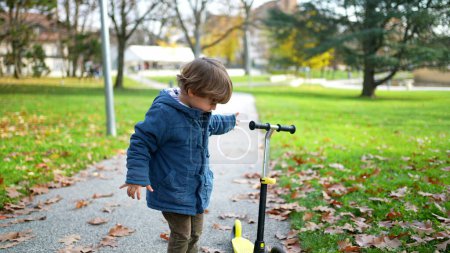 Téléchargez les photos : Un petit garçon debout au parc pendant la journée d'automne portant une veste bleue vérifiant son scooter à 3 roues - en image libre de droit