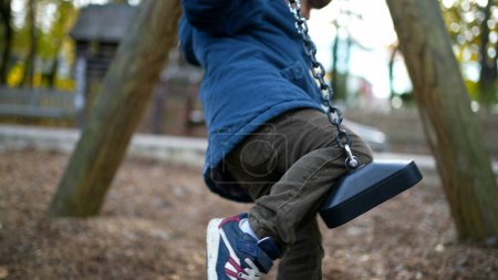 Photo for Child trying to climb park swing during autumn day wearing blue jacket. One small 3 year old boy attempts to sit on top of swing - trial and error childhood concept - Royalty Free Image
