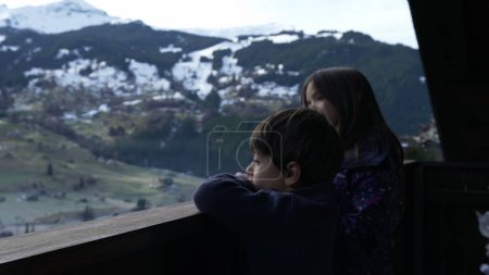 Photo for Young brother and sister standing in wooden Swiss chalet balcony overlooking mountains covered in snow during winter ski season, children enjoying Alps landscape - Royalty Free Image