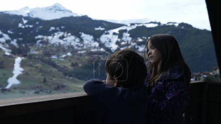 Téléchargez les photos : Jeune frère et sœur debout dans un chalet suisse en bois balcon donnant sur les montagnes couvertes de neige pendant la saison de ski d'hiver, enfants profitant du paysage des Alpes - en image libre de droit