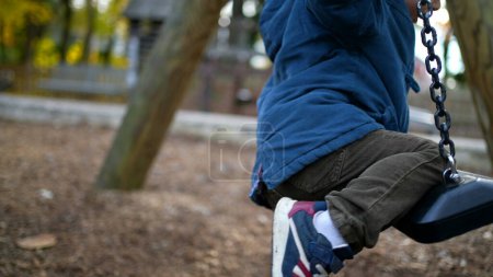 Photo for Child trying to climb park swing during autumn day wearing blue jacket. One small 3 year old boy attempts to sit on top of swing - trial and error childhood concept - Royalty Free Image