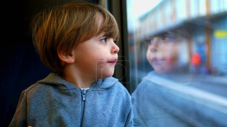 Téléchargez les photos : Un petit garçon voyageant en train penché sur le verre regardant fixement, reflet du visage de l'enfant sur la fenêtre - en image libre de droit