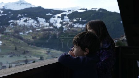 Photo for Young brother and sister standing in wooden Swiss chalet balcony overlooking mountains covered in snow during winter ski season, children enjoying Alps landscape - Royalty Free Image