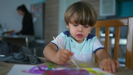 Foto de El niño artístico juega con la pintura de acuarela en casa, mientras que la madre hace las tareas de fondo. Pintura creativa de niño con pincel - Imagen libre de derechos