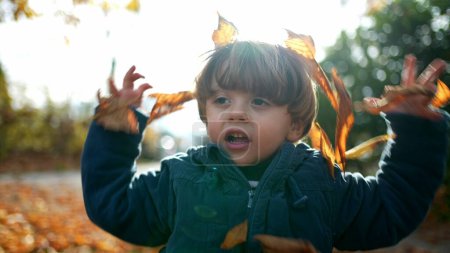 Téléchargez les photos : Enfant avec des feuilles orange sur la tête dans Sunlit Park - Automne Playtime of kid wearing blue jacket amidst orange nature playing outdoors - en image libre de droit