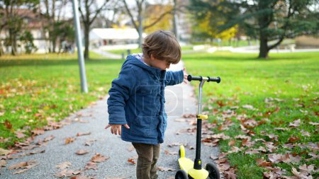 Photo for One little boy standing at park during autumn day wearing blue jacket checking his 3 wheeled scooter - Royalty Free Image