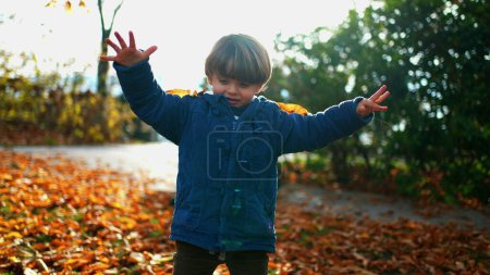 Photo for Playful child putting orange leaves in head while standing out sunlit park during autumn fall season. 3 year old kid plays by himself in nature - Royalty Free Image
